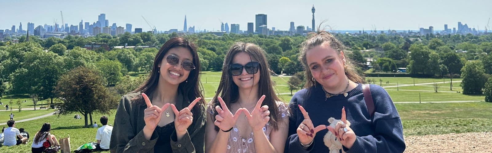 Three students make a "W" with their hands while standing in front of the London skyline